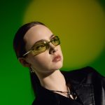 Junge Frau mit sportlicher Y2K-Sonnenbrille und schwarzem Oberteil vor grün beleuchtetem Hintergrund